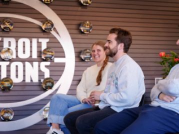 Three Pistons' employees giving a Q and A sit on high chairs with the Detroit Pistons logo on the wall in the background
